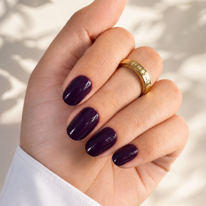 Hand with dark purple nail polish wearing a gold ring on a light background