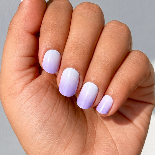Hand with light purple to white ombre nails on a neutral background