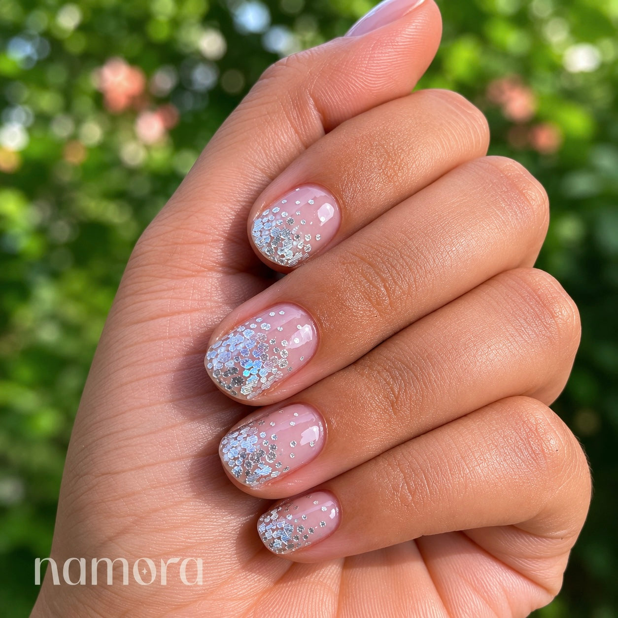 Hand with glittery silver namora nail wraps against a blurred green background