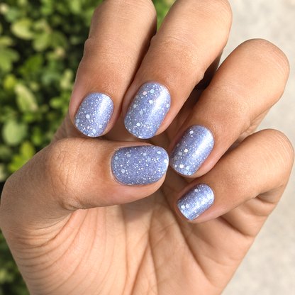Hand with light blue glittery nail wraps by namora against a blurred green background