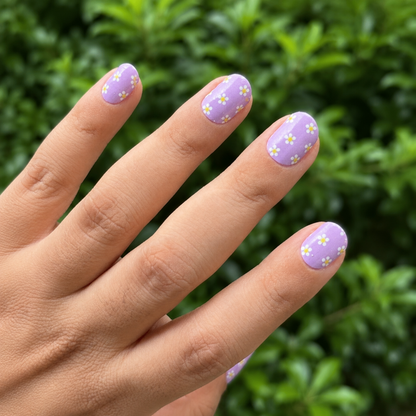 Hand with purple nail wraps featuring white floral designs against a green leafy background