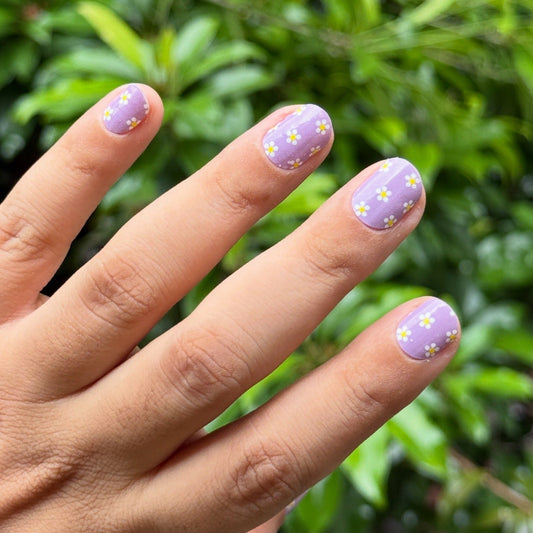 Hand with plavender nail wraps featuring white floral designs against a green leafy background