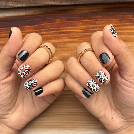 Two hands with black and white leopard print nail wraps on a wooden surface.