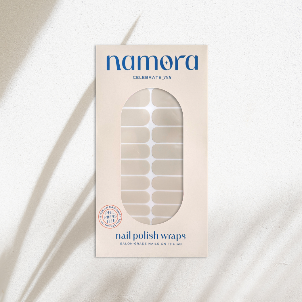A package of Namora nail polish wraps in a sheer miilky white shade, shown in its cream-colored box with a clear window displaying the nail wraps inside. The packaging features blue text and is set against a textured white background with soft, diagonal shadows.