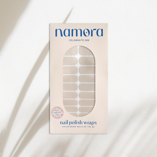 A package of Namora nail polish wraps in a sheer miilky white shade, shown in its cream-colored box with a clear window displaying the nail wraps inside. The packaging features blue text and is set against a textured white background with soft, diagonal shadows.